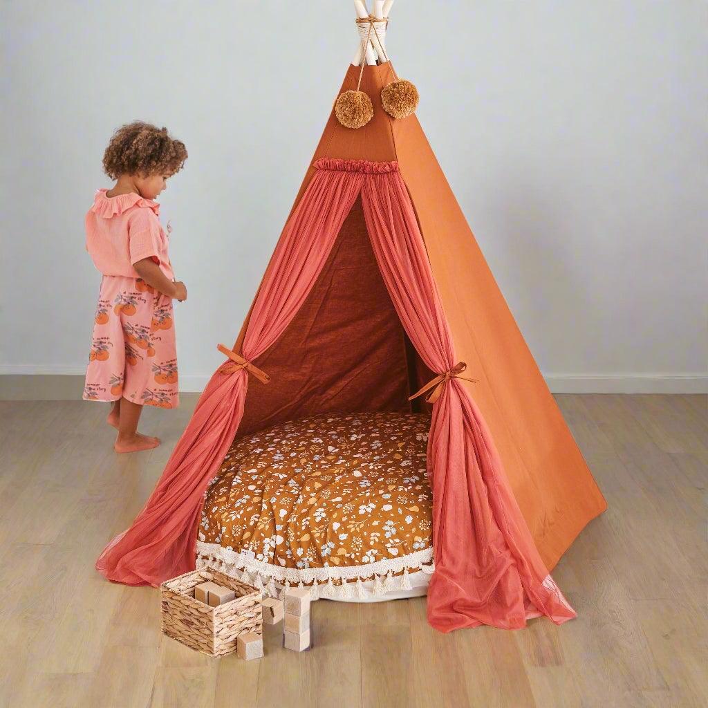 Child stood next to an orange and pink teepee with a brown patterned floor cushion inside