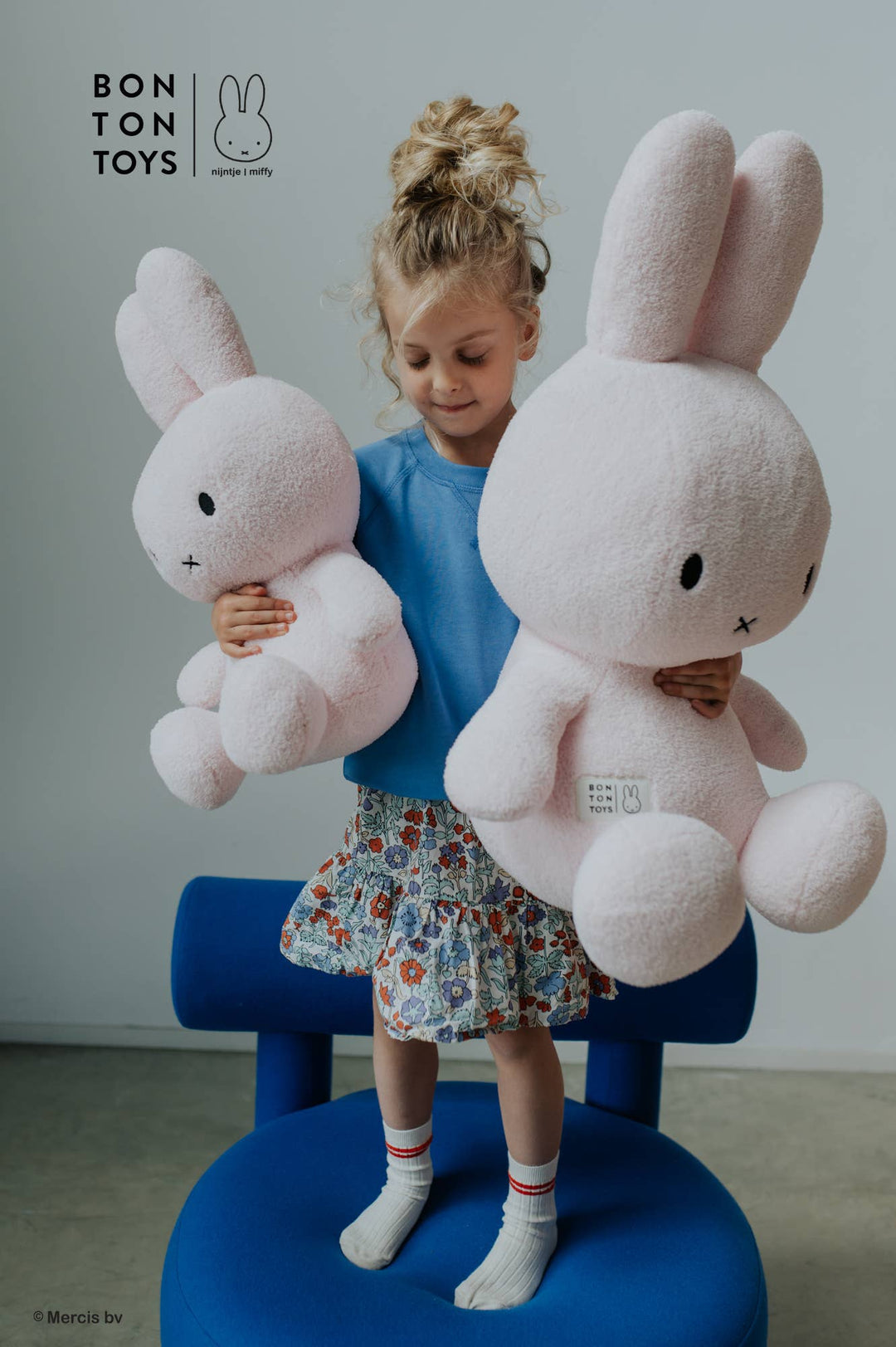 Child holding two large pink plush toys bunnies, with Bonton Toys logo above.