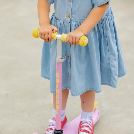 Child in a blue dress riding a pink scooter with yellow handlebars on a neutral background
