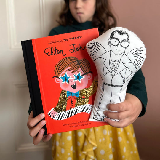 A child holding a cushion doll with a screen printed design of Elton John in black and white, and a book with Elton John's face on the cover.