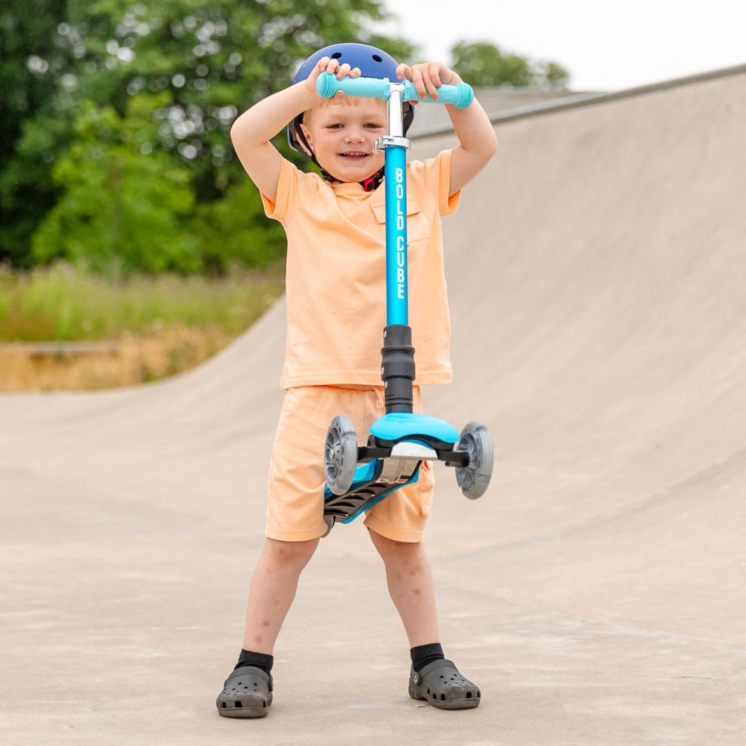 Child with a blue scooter on a paved path with greenery in the background