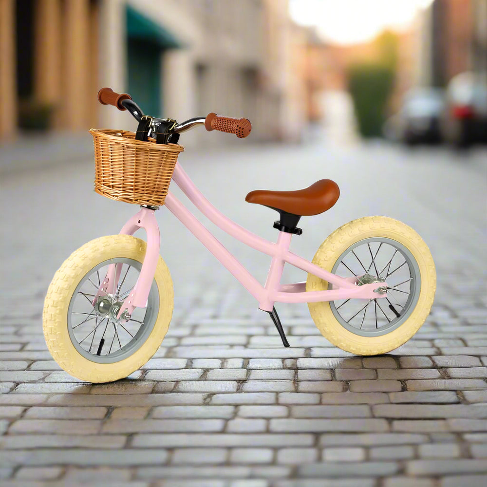 Balance bike with pale pink metal work and cream wheels angled to the side with a brown saddle and brown handles with a brown wicker basket attached to the handlebar