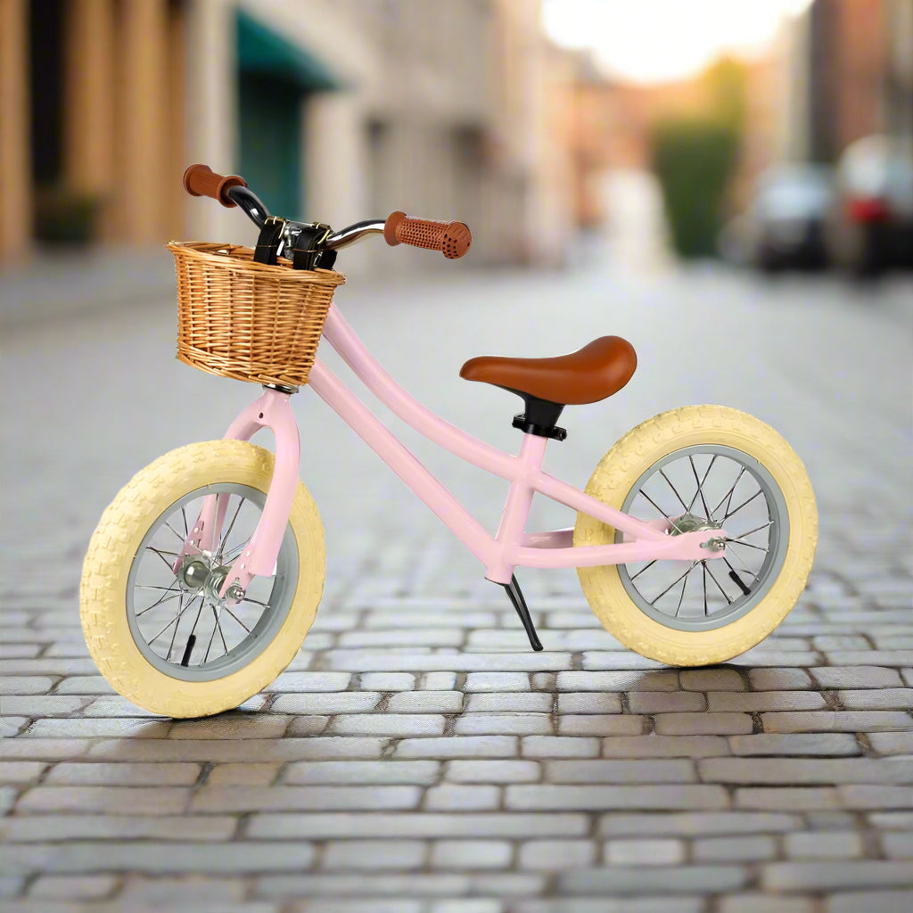 Balance bike with pale pink metal work and cream wheels angled to the side with a brown saddle and brown handles with a brown wicker basket attached to the handlebar