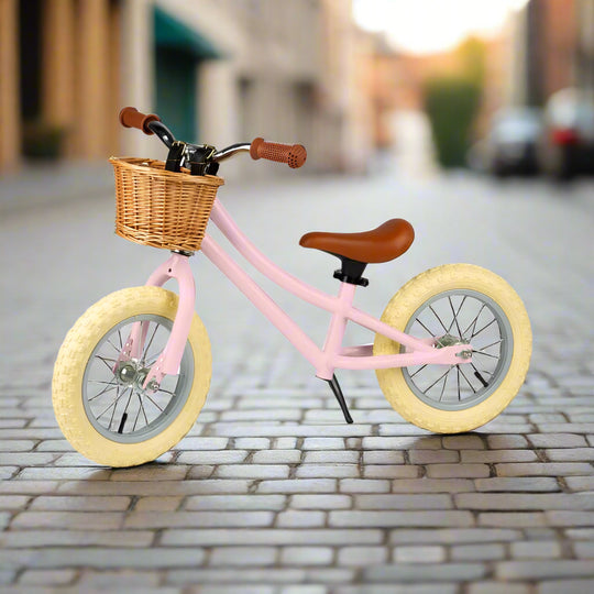 Balance bike with pale pink metal work and cream wheels angled to the side with a brown saddle and brown handles with a brown wicker basket attached to the handlebar