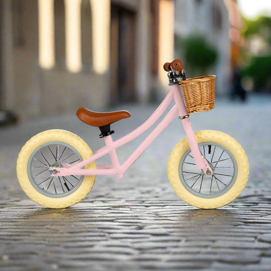 Balance bike with sage green metal work and cream wheels with a brown saddle and brown handles with a brown wicker basket attached to the handlebar