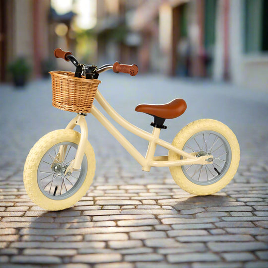 Cream balance bike to the side, showing cream coloured metal work and cream wheels, with a brown seat and handles, and a crown wicker basket attached to the handlebar