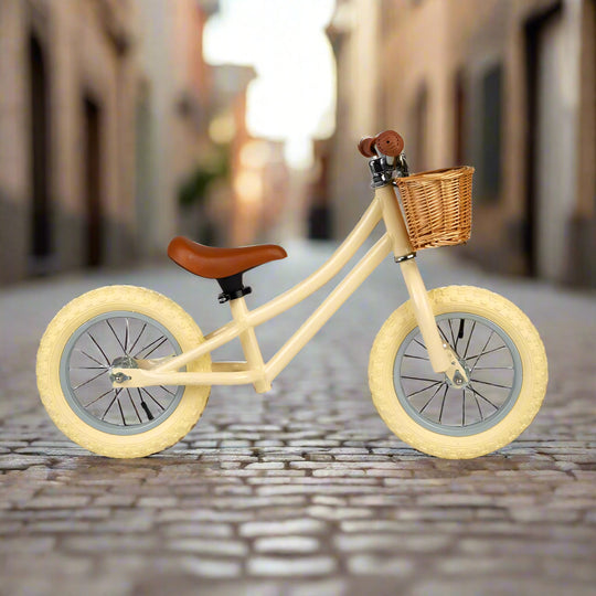 Cream balance bike to the side at a different angle, showing cream coloured metal work and cream wheels, with a brown seat and handles, and a crown wicker basket attached to the handlebar