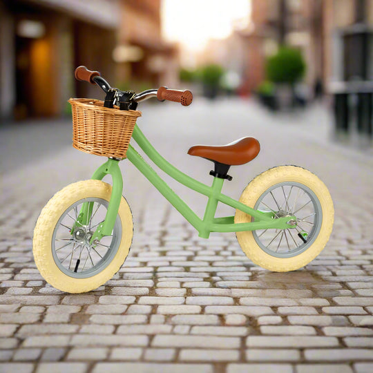 Balance bike with sage green metal work and cream wheels angled to the side with a brown saddle and brown handles with a brown wicker basket attached to the handlebar