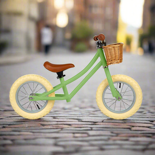Balance bike with sage green metal work and cream wheels angled to the side with a brown saddle and brown handles with a brown wicker basket attached to the handlebar