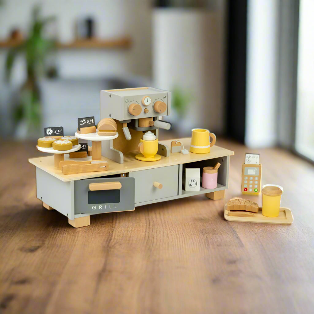 Wooden pretend play coffee machine on a play counter with a grill, cupboard, cake plates with tongs, a frothy coffee in the coffee maker, and a tray with a wooden pastry and a takeaway coffee cup. All made of wood