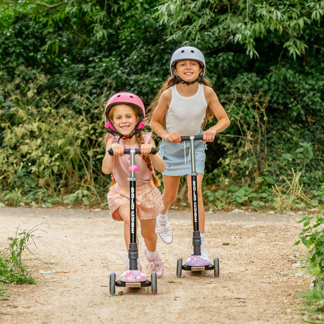 Two children riding scooters outdoors with greenery in the background