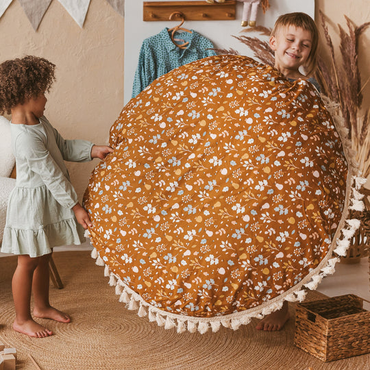 Two children holding a round floral-patterned floor cushion in a room with a wooden floor and decorative elements.