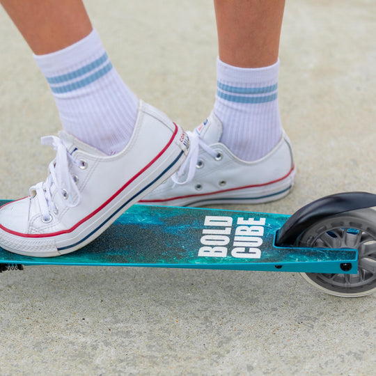 Person wearing white trainers with red accents on a teal scooter with 'BOLD CUBE' branding.
