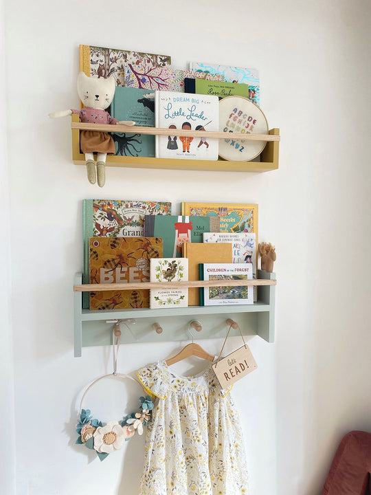 Two bookshelves, one on top of the other on a wall, the bottom one has peg rails and things hanging from it. Both bookshelves are full of children's books and toys