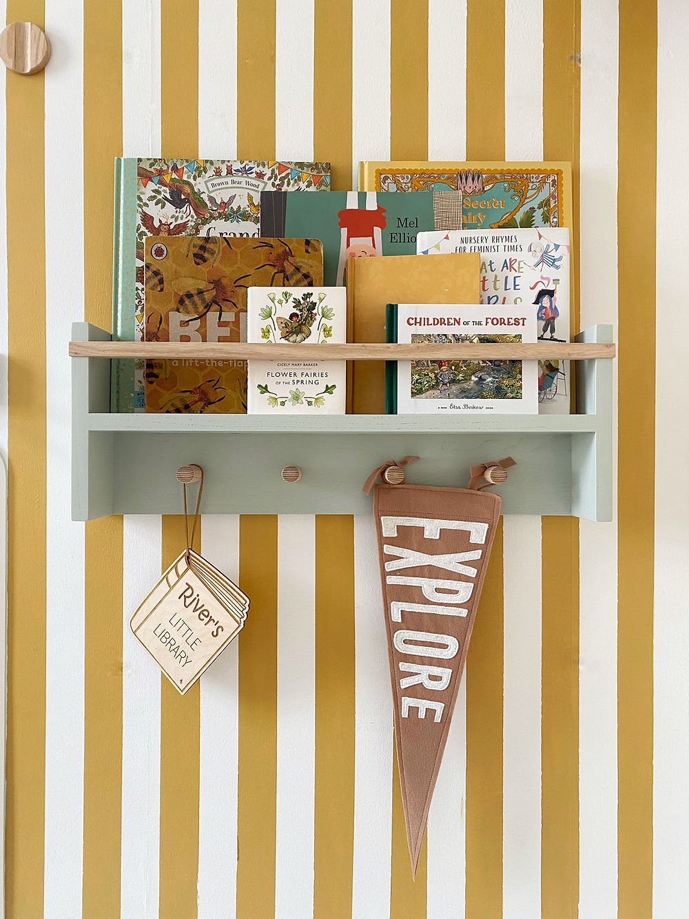 A wall-mounted peg rail bookshelf in a room with striped yellow and white wallpaper, displaying various children's books and decorative items.