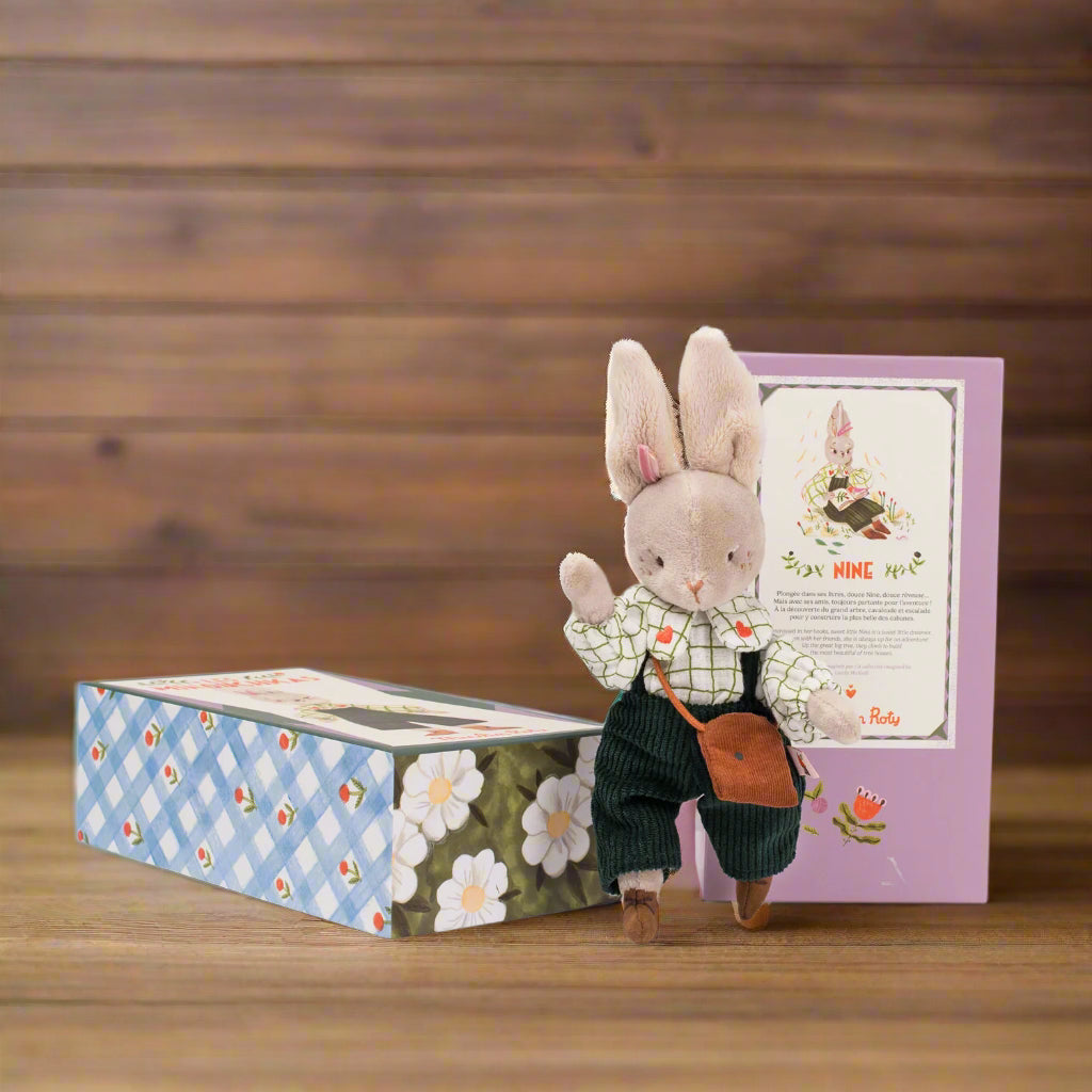 Stuffed rabbit toy with a white checkered shirt and navy corduroy dungarees standing next to a decorative box on a white background