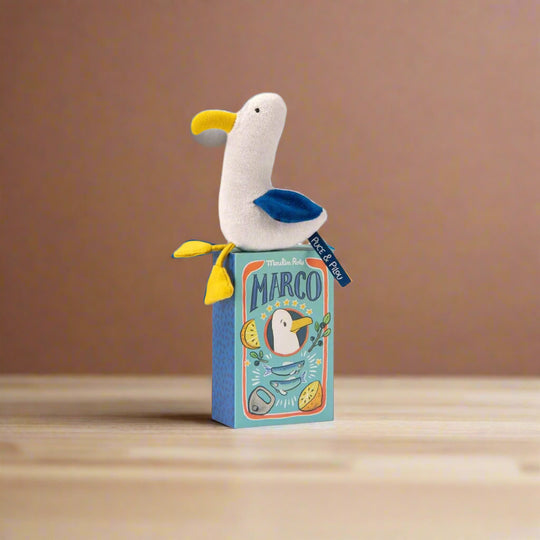 Plush seagull toy rattle with white body, yellow beak and feet and blue wings sitting on its decorative packaging box