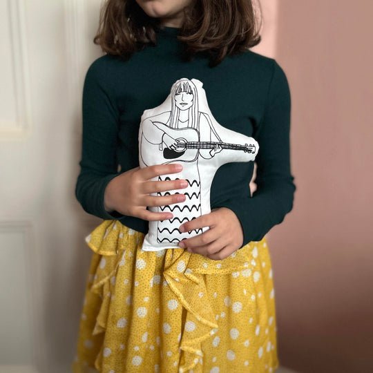 A child holding screen printed doll cushion of singer-songwriter Joni Mitchell playing a guitar