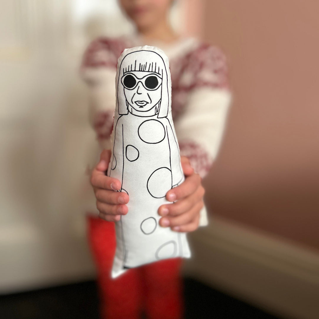 A child holding a screen printed cushion doll with a white front featuring a black outlined drawing and polka dots, on a blurred background.
