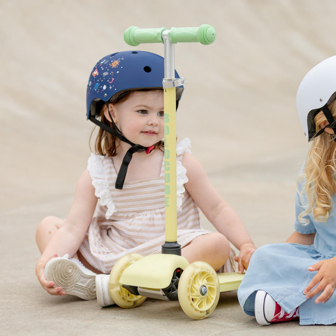 Two children sitting on the floor with balance scooters and helmets.