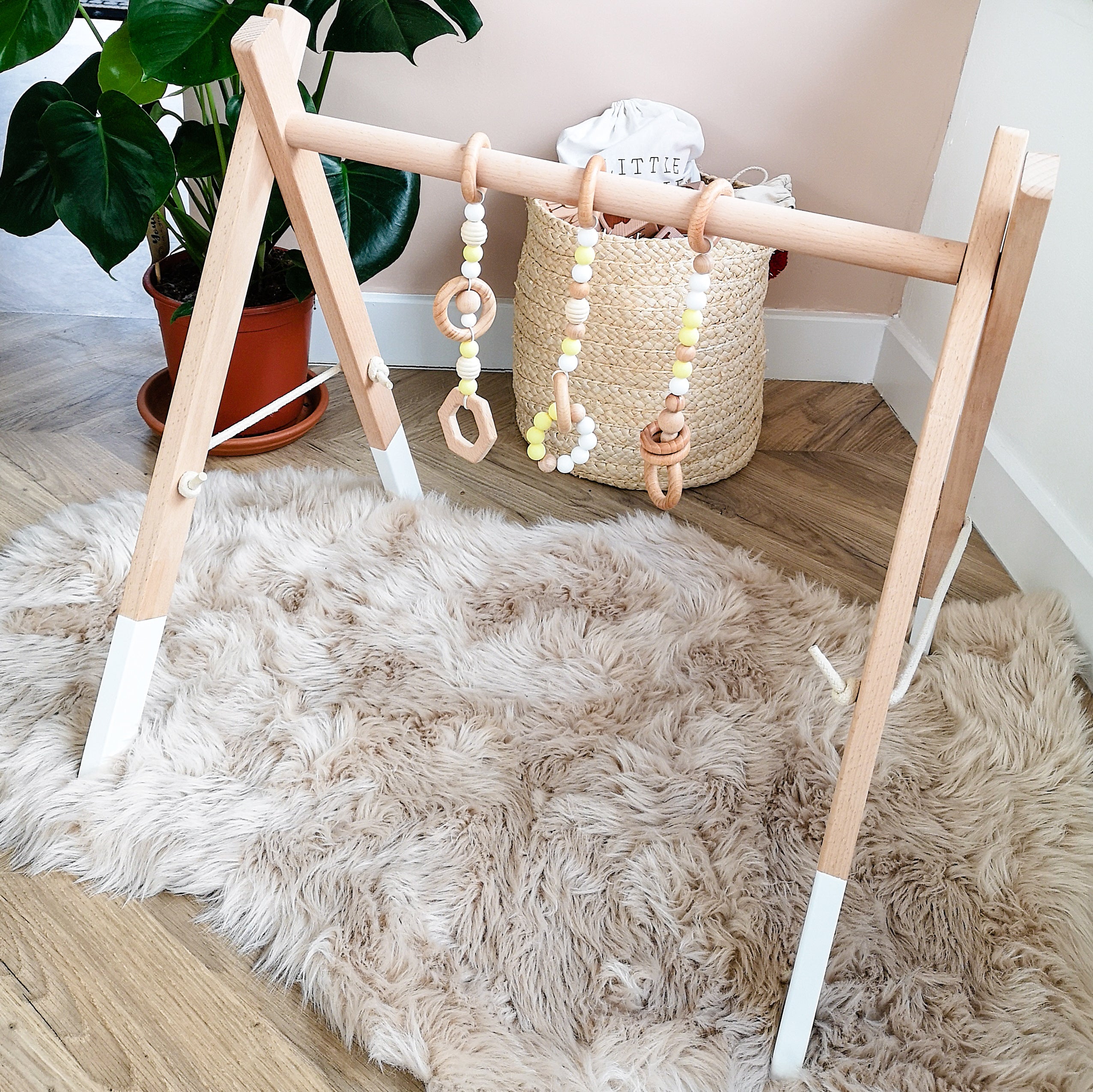 A beech wood play gym with yellow accents is set up on a furry rug, featuring a wooden frame and hanging wooden toys.