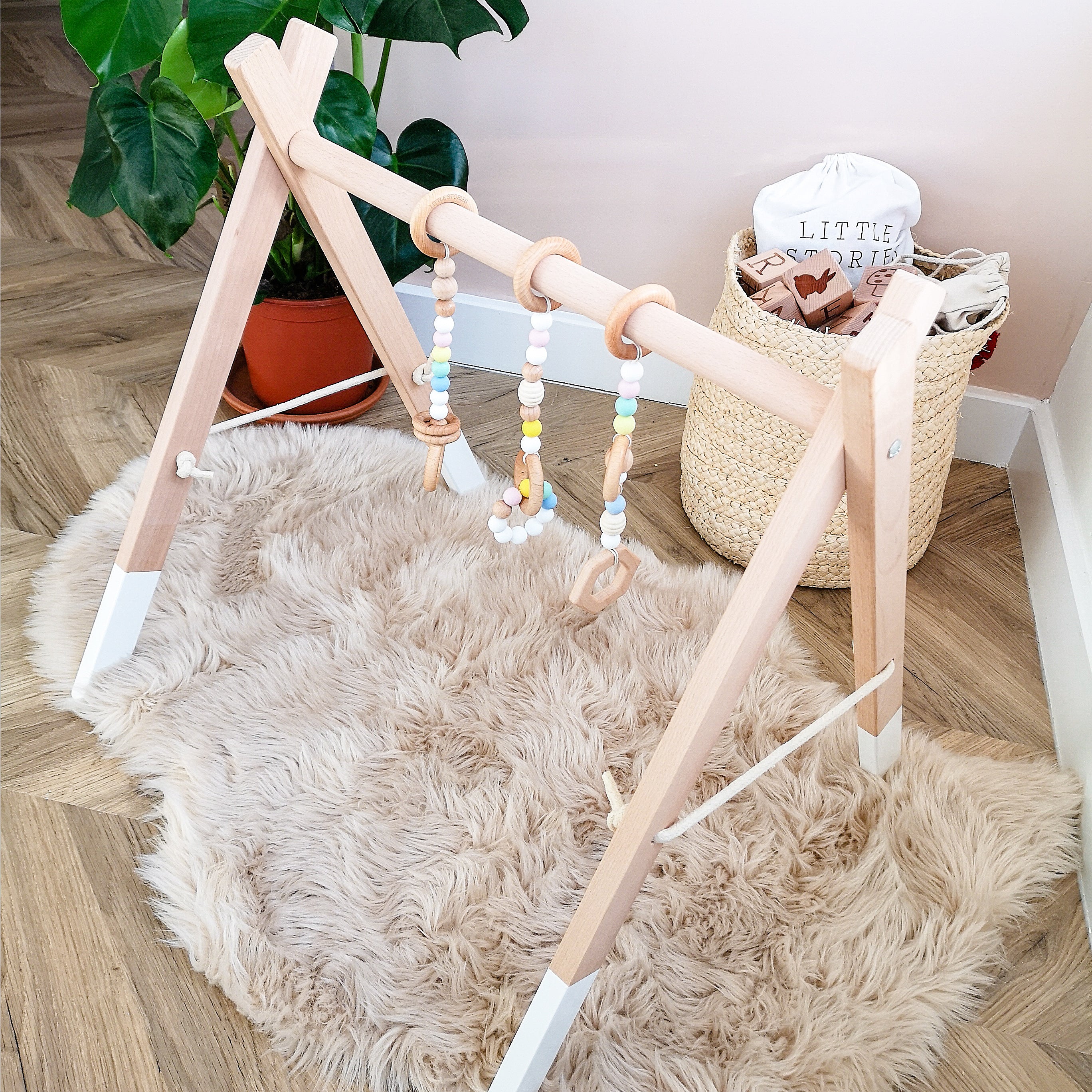 A beech wood baby play gym with pastel coloured hanging toys, set up on a furry rug in a room.
