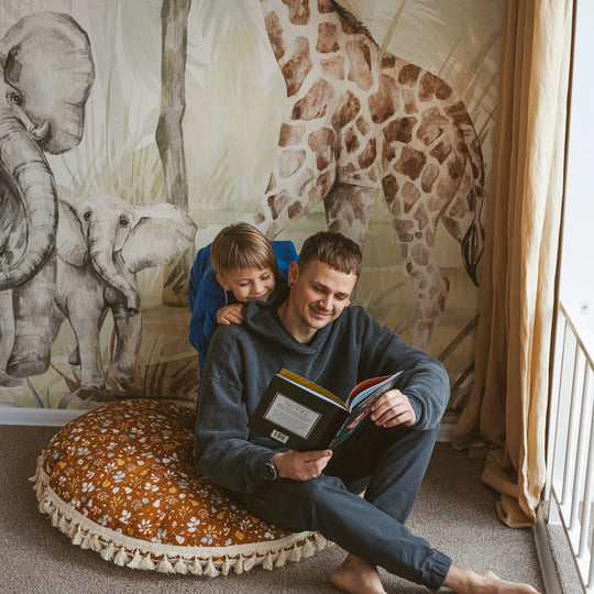 Man and child sitting on a round cushion in front of a wall with animal murals.