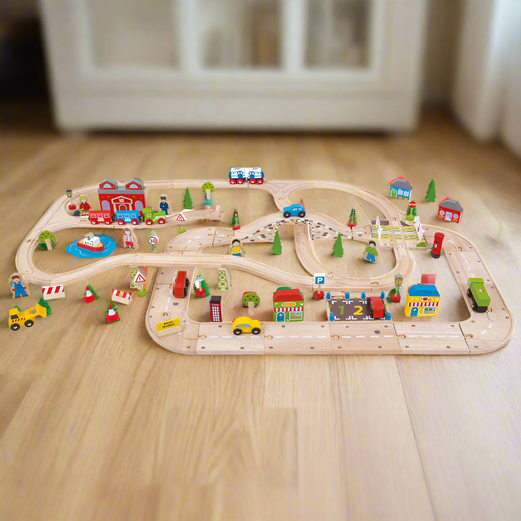 Extensive wooden train set depicting a city road and railway network with various play pieces including cars, trains, tracks, buildings, and a boat.