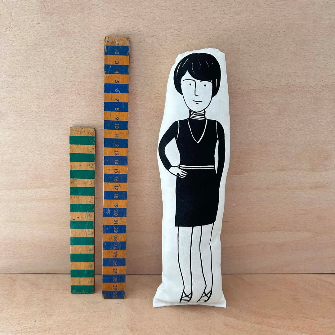 Screen printed doll cushion of Bridget Riley next to two wooden rulers on a wooden background
