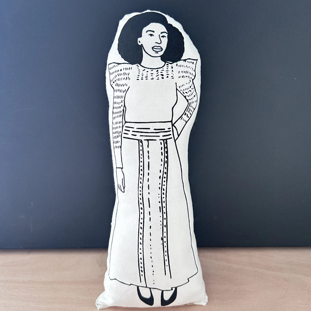 Screen printed doll cushion with white fabric and black illustration depicting author, feminist and TED speaker Chimamanda Ngozi Adichie wearing a long traditional dress with one hand on her hip