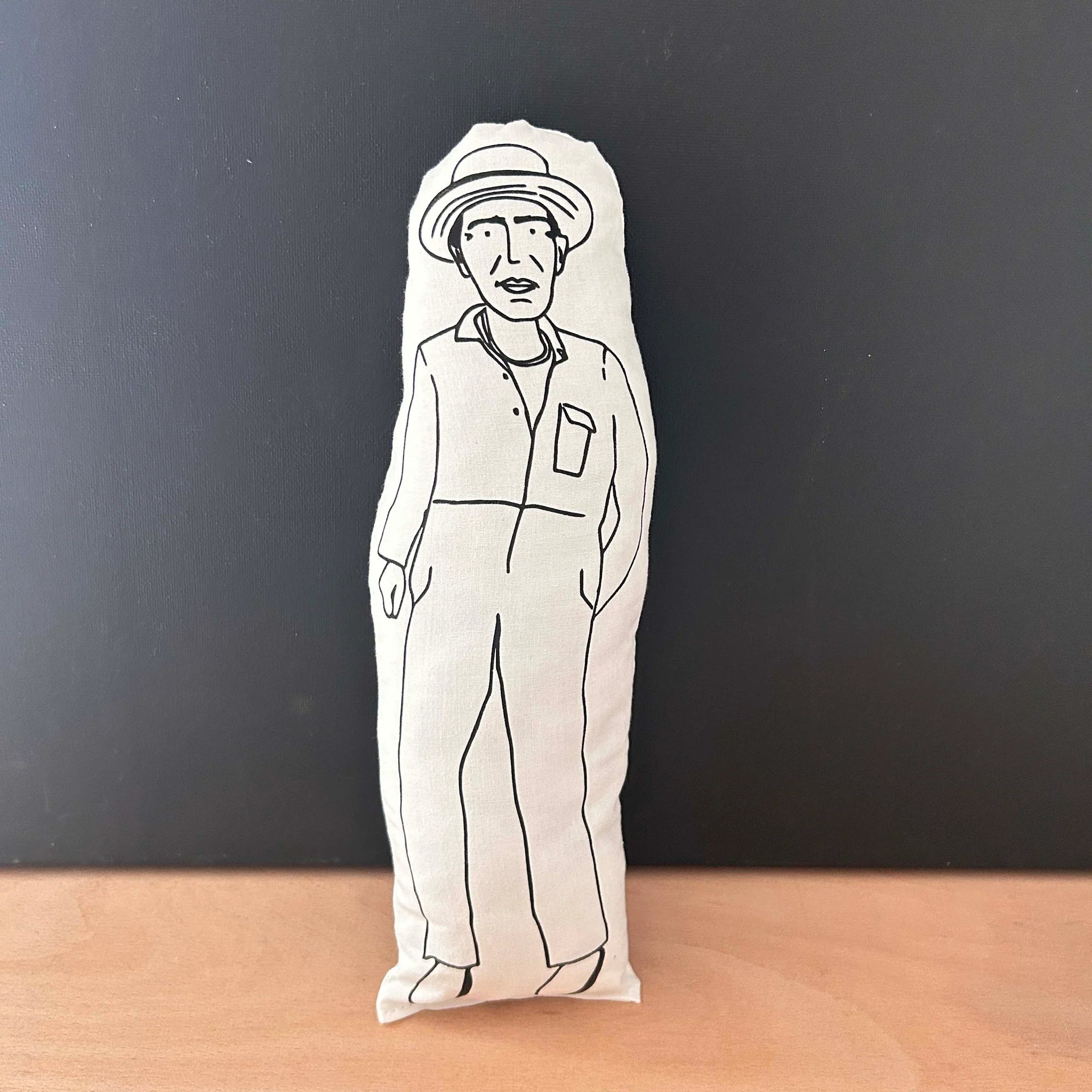 Screen printed doll cushion with white fabric and black illustration depicting filmmaker Derek Jarman wearing gardening overalls and a straw ha