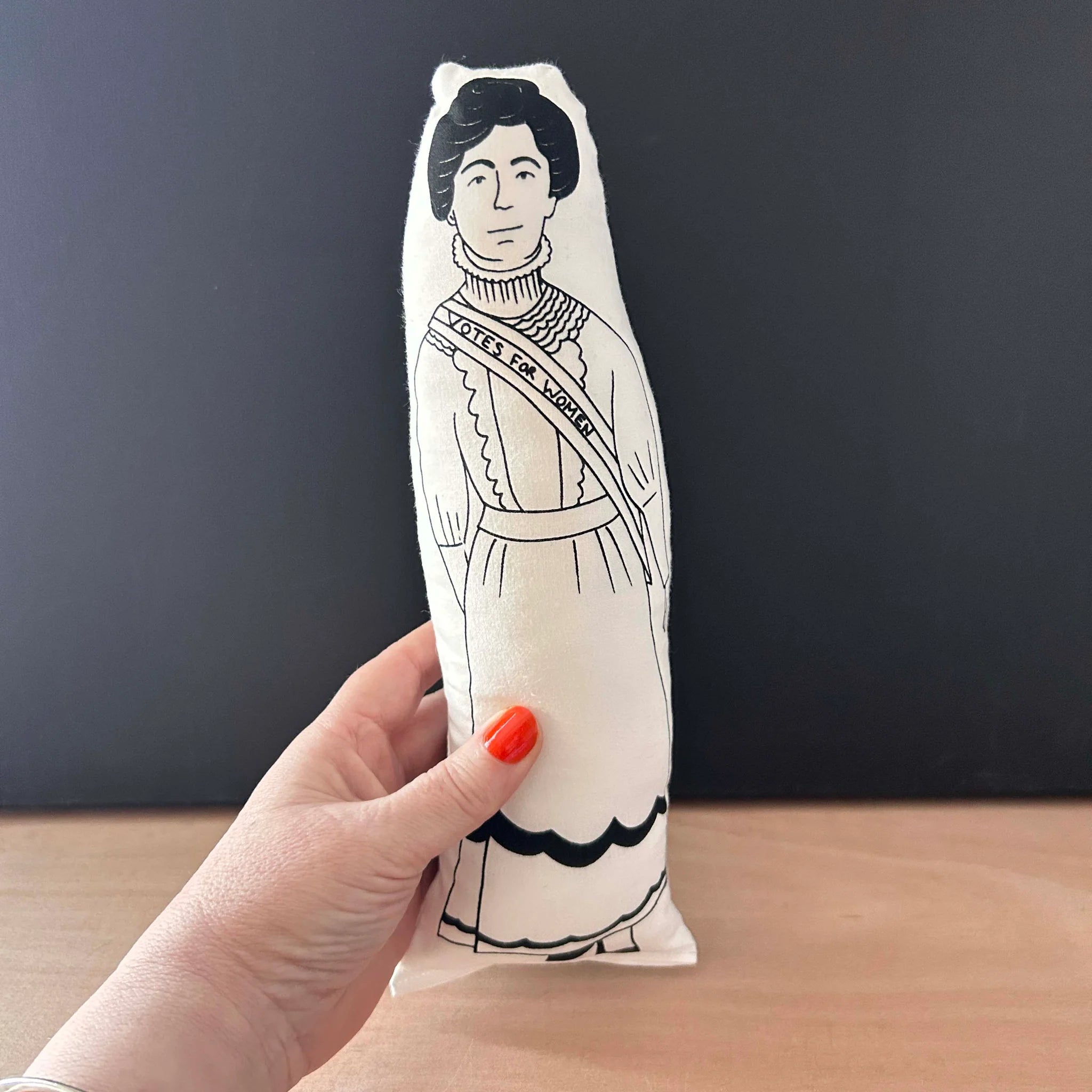 A hand holding a screen printed doll cushion with white fabric and black illustration depicting women's rights activist Emmeline Pankhurt, wearing Edwardian dress with a sash declaring 'votes for women'