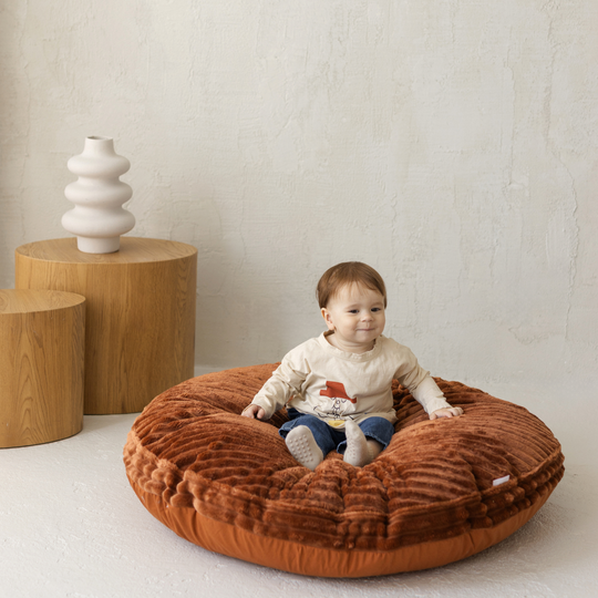 Child sitting on a brown plush cushion in a minimalistic room with wooden tables and a white vase.
