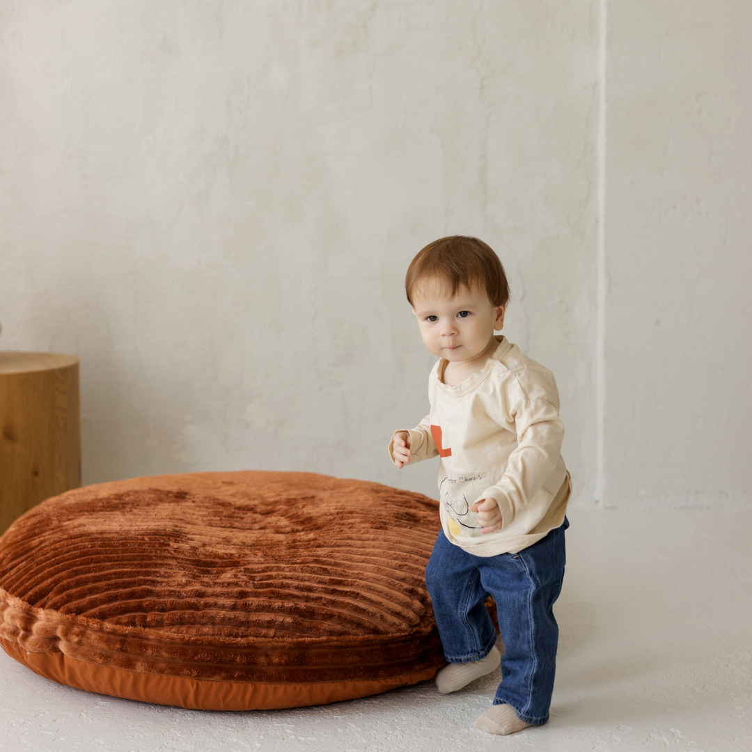 Child standing next to a large brown round cushion on a light floor.
