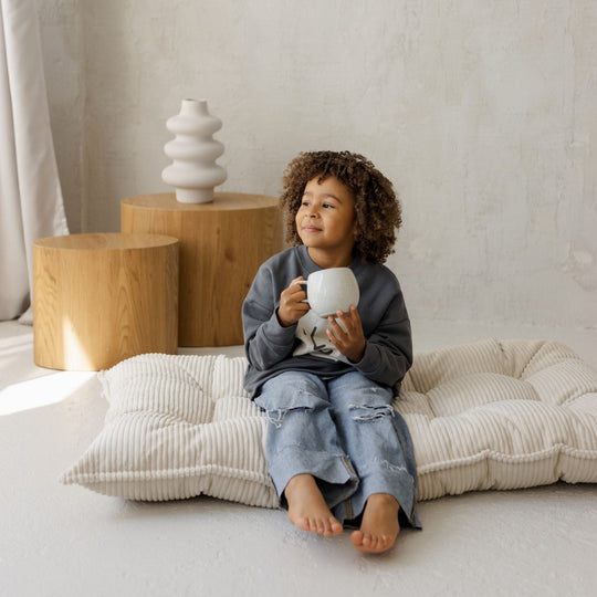 Child sitting on a cushion holding a white mug in a minimalistic room with wooden tables and decorative items.