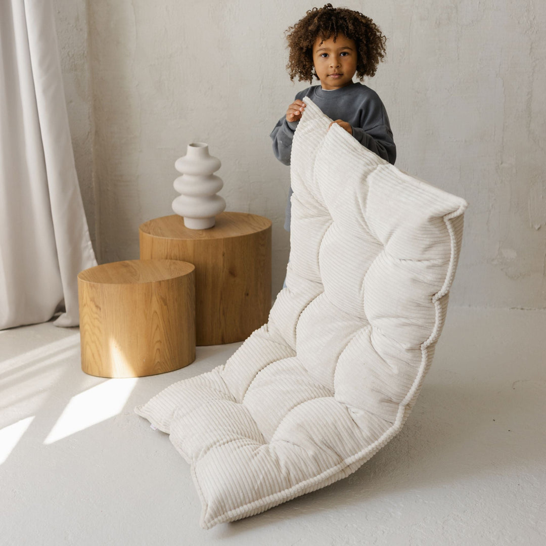 Child holding a large white cushion in a minimalistic room with wooden stools and a vase.