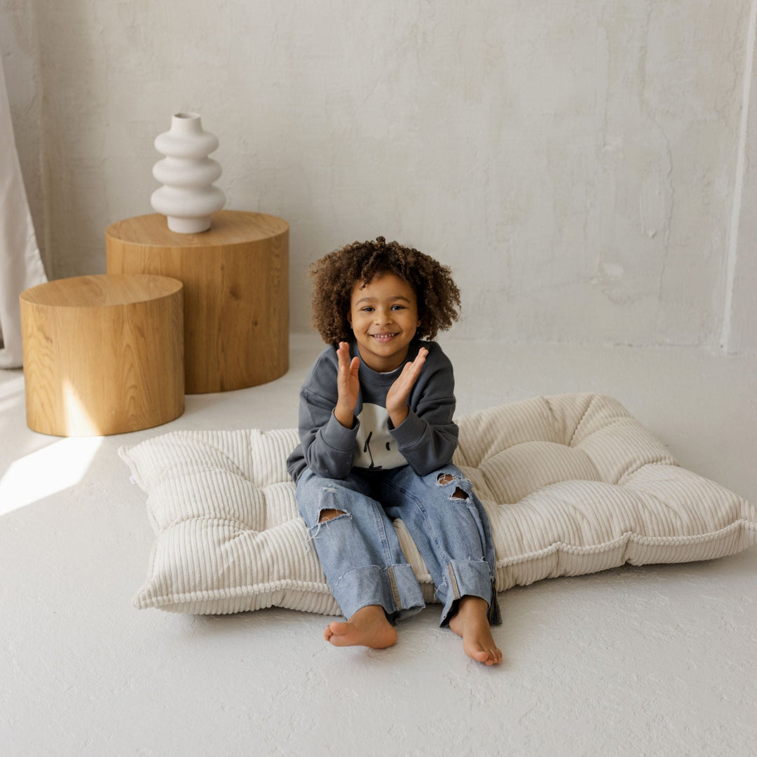 Child sitting on a large cushion in a minimalistic room with wooden stools and a vase.
