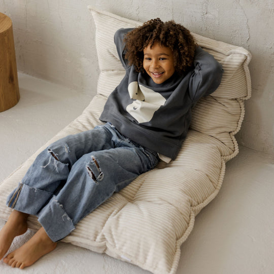 Child sitting on a textured beige pillow folded so his back is resting on the pillow against the wall