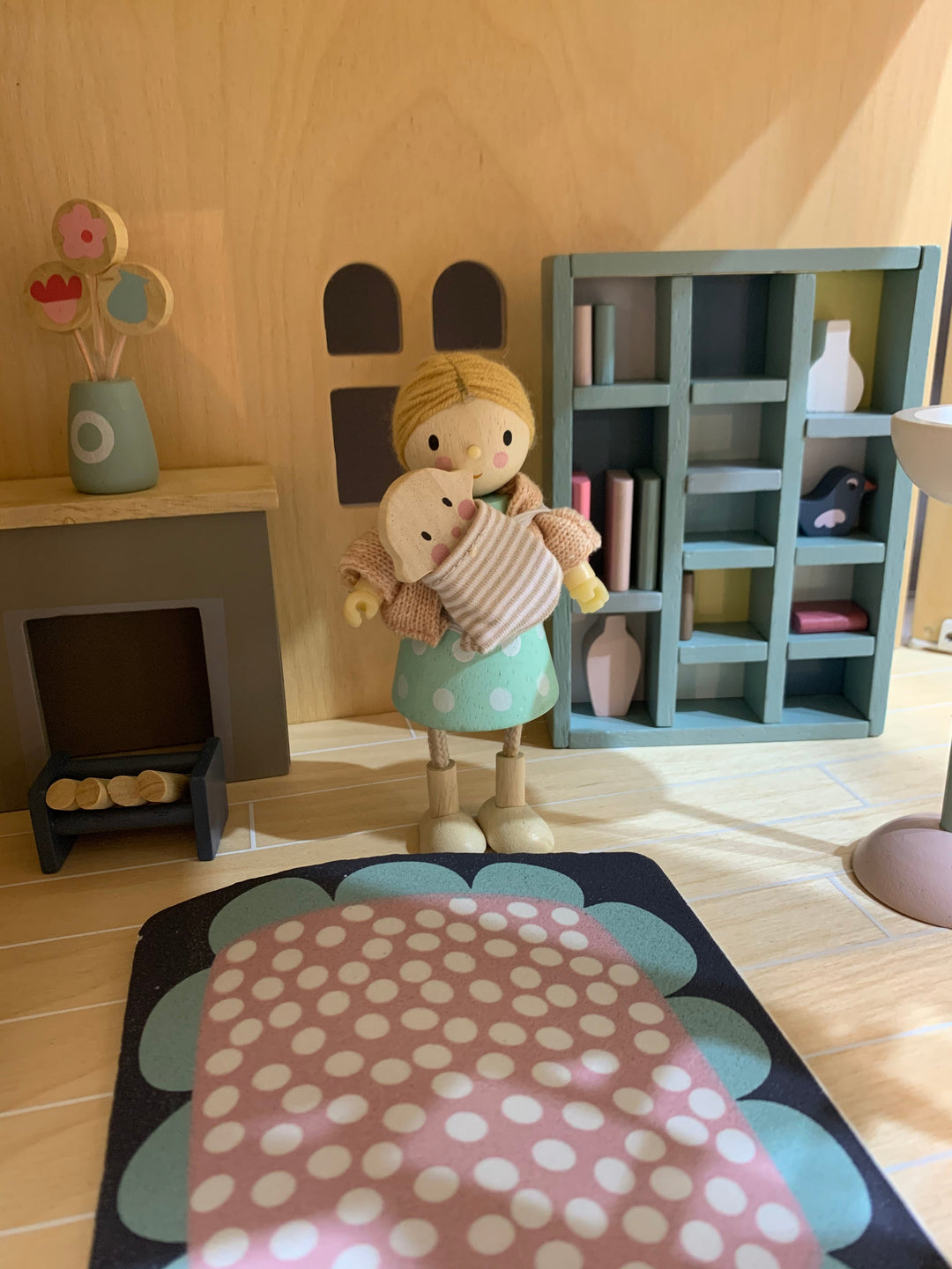 Toy figure of a girl holding a teddy bear in a miniature room setting with shelves and fireplace.