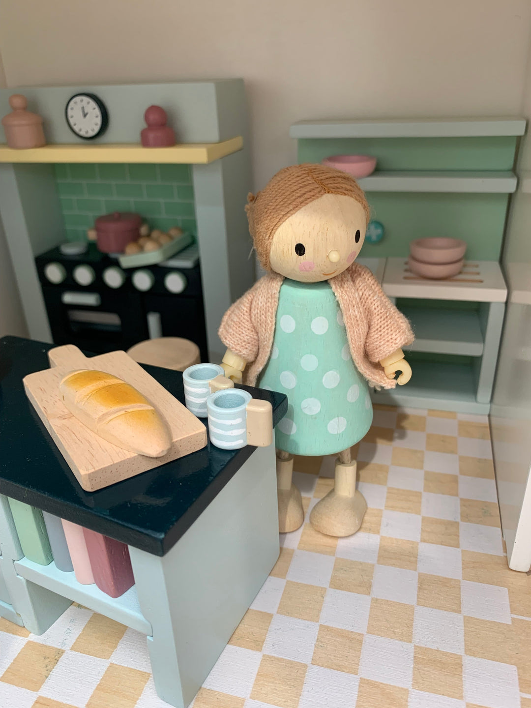 Toy kitchen set with a doll in a green dress and beige cardigan standing near a counter with bread and cups.