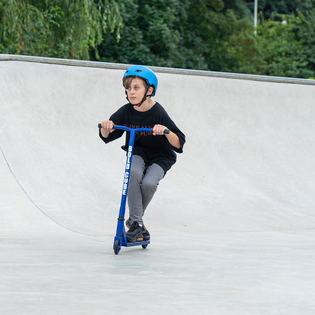 Child riding a blue scooter at a skate park with greenery in the background