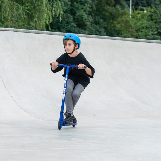 Child riding a blue scooter at a skate park with greenery in the background