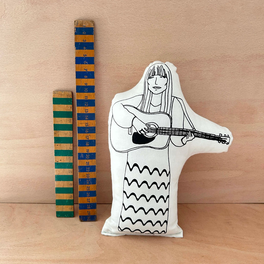 Screen printed doll cushion of singer-songwriter Joni Mitchell playing a guitar next to two wooden rulers