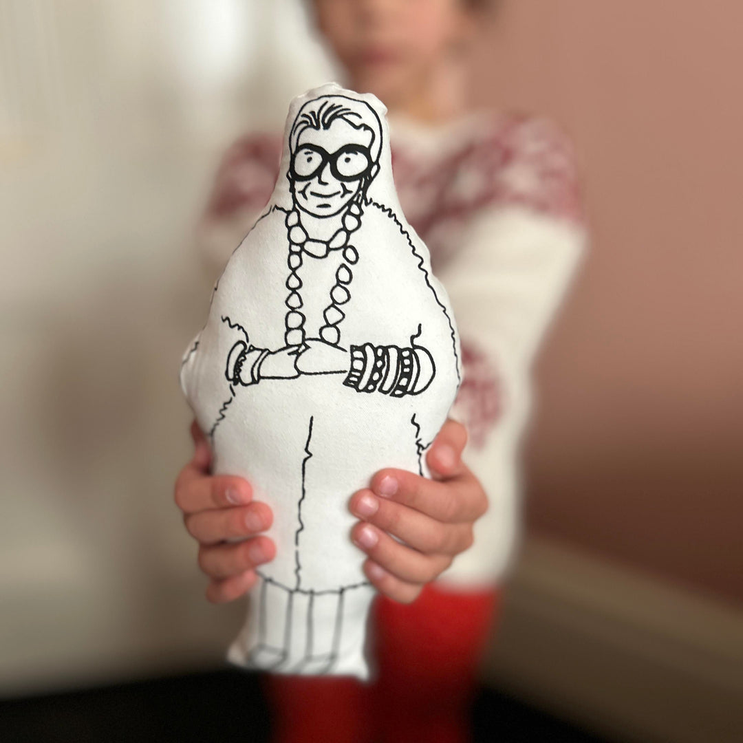 A child holding out screen printed doll cushion of Iris Apfel towards the camera with a blurred background