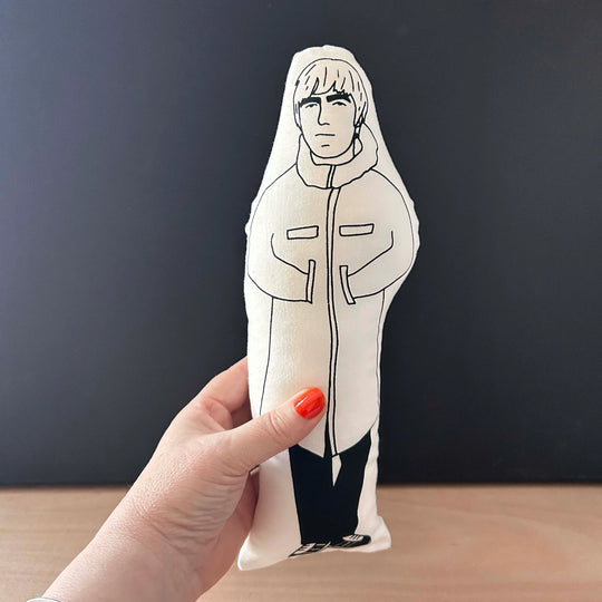 Hand holding screen printed doll cushion with white fabric and black illustration depicting Liam Gallagher wearing a large coat with his hands in his pockets