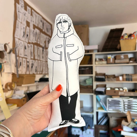 A hand holding  screen printed doll cushion of Liam Gallagher against an out of focus interior setting