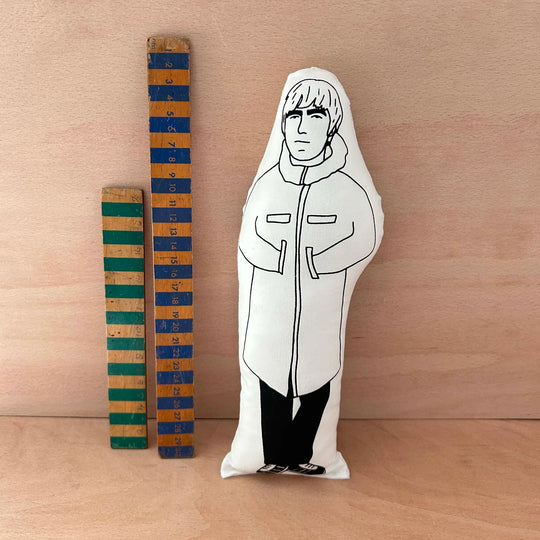 Screen printed doll cushion of Liam Gallagher next to two wooden rulers on a wooden background