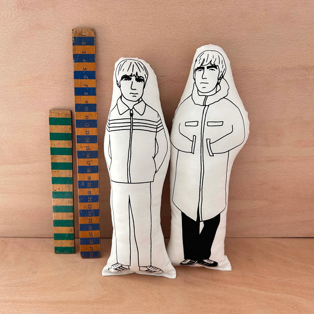 Two screen printed doll cushions, one of Liam Gallagher and one of his brother Noel Gallagher, together making the band Oasis, next to two wooden rulers on a wooden background
