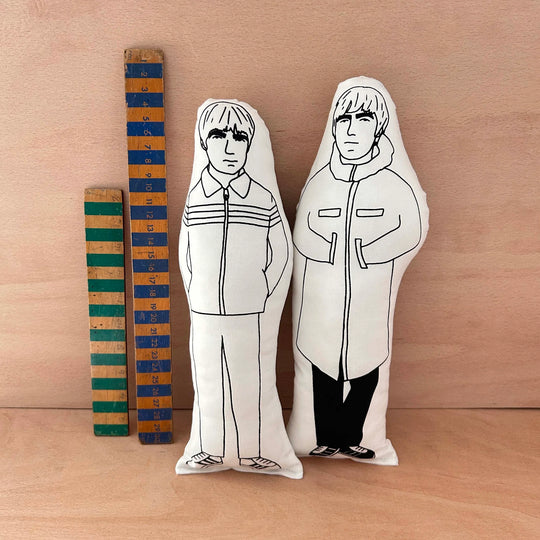Two screen printed doll cushions, one of Liam Gallagher and one of his brother Noel Gallagher, together making the band Oasis, next to two wooden rulers on a wooden background