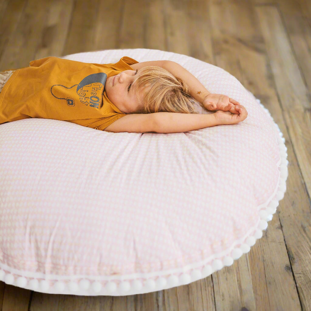 Chil lying on his back on a large pale pink floor cushion with his hands above his head